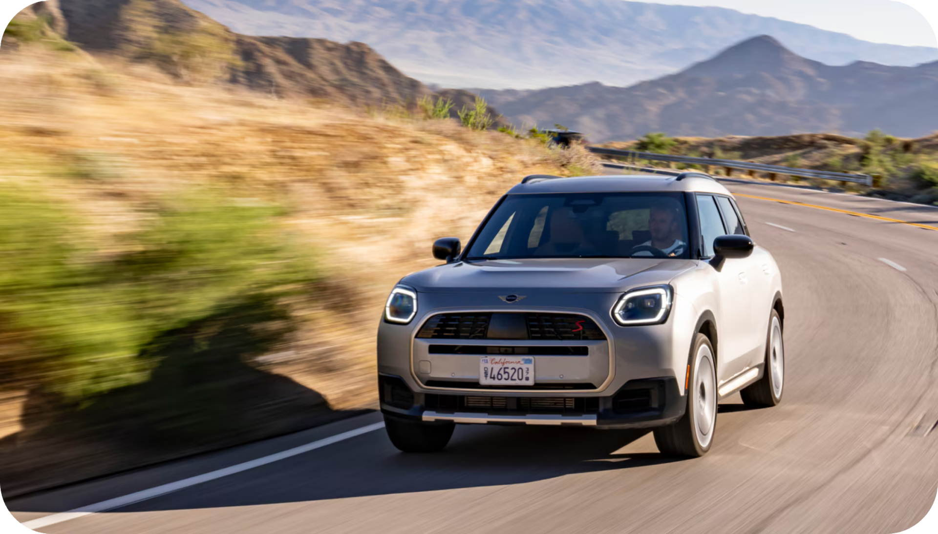 A front-view of a 2025 MINI Countryman S ALL4 turning around a bend with a blurred mountainous terrain in the background.
